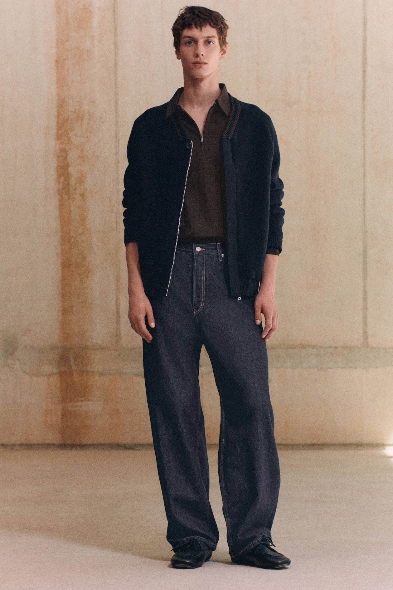 A person in a black jacket, brown shirt, and dark jeans stands against a textured beige wall.