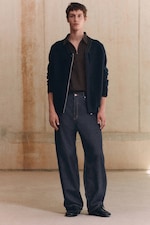 Placeholder for: A person in a black jacket, brown shirt, and dark jeans stands against a textured beige wall.