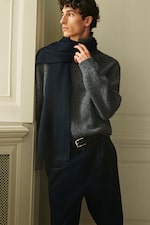 Placeholder for: A person wearing a gray sweater and dark scarf leans against a wall, looking to the side with a thoughtful expression.