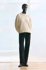 Placeholder for: Person in a cream sweater and dark pants stands in front of a window with hands in pockets.