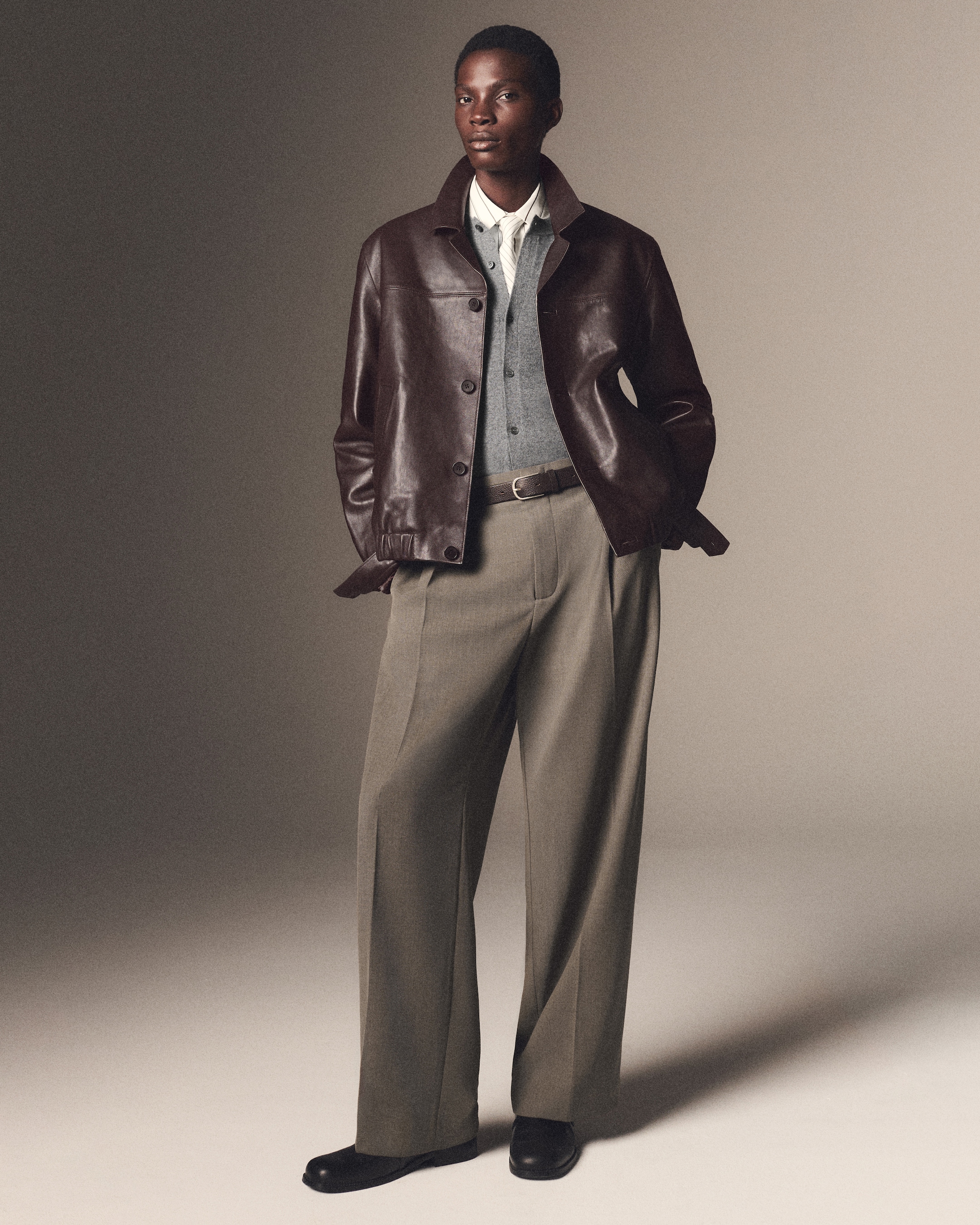 A person wearing a brown leather jacket over a gray sweater and white shirt, paired with tan wide-leg trousers and black shoes, standing confidently.