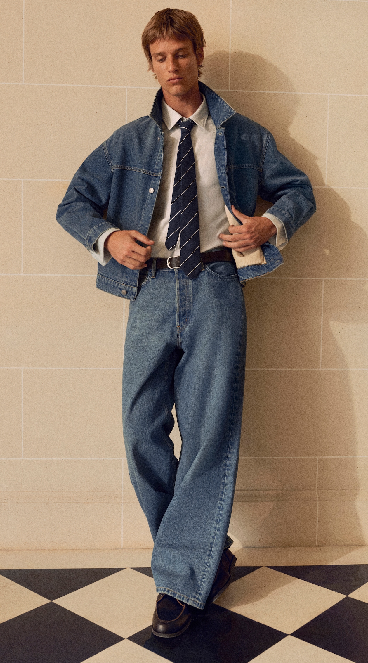 Person wearing a denim jacket, wide-leg jeans, white shirt, and striped tie, standing on a checkered floor against a tiled wall.