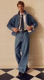 Placeholder for: Person wearing a denim jacket, wide-leg jeans, white shirt, and striped tie, standing on a checkered floor against a tiled wall.