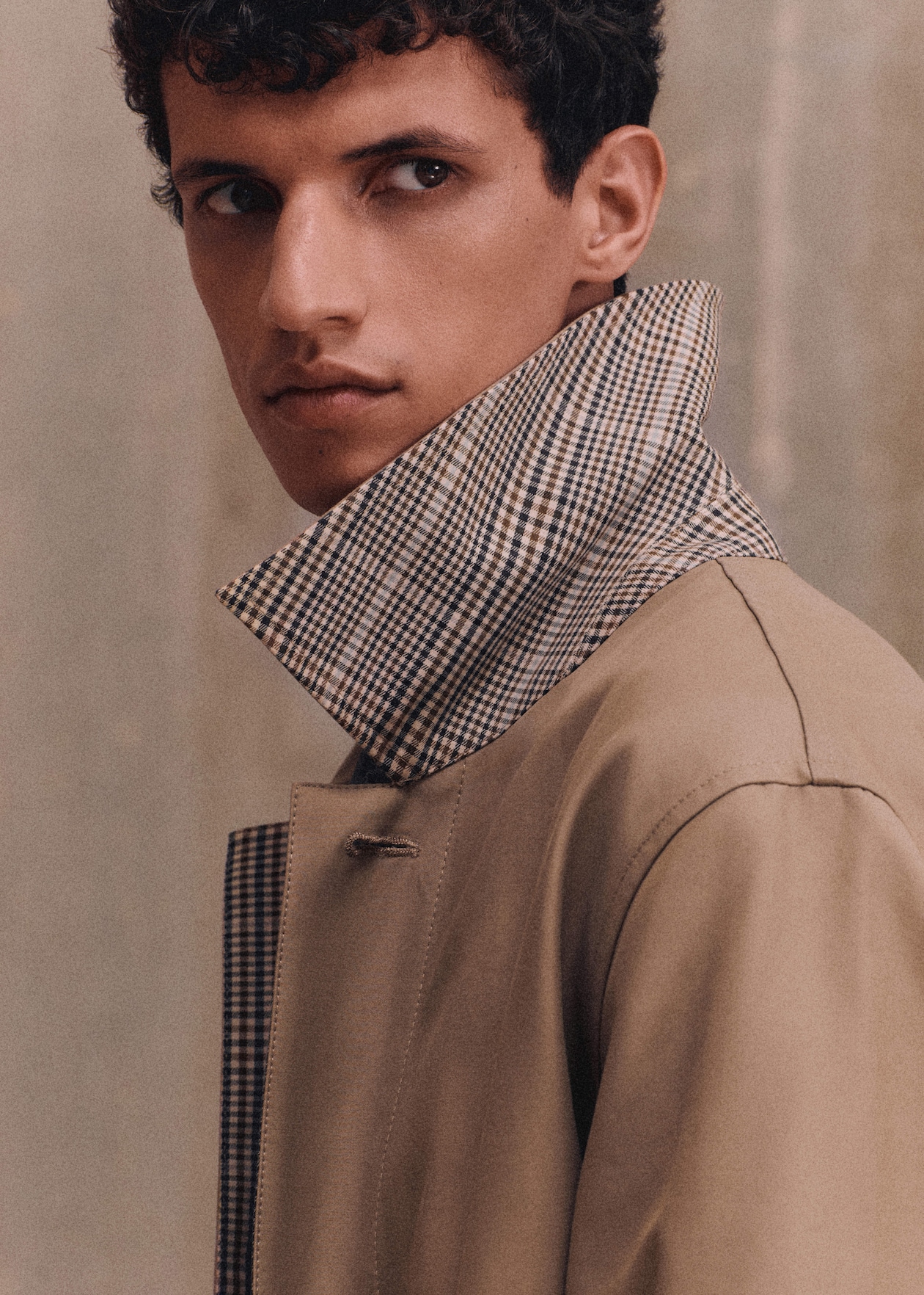 A person with curly hair looking to the side, wearing a beige coat with a checkered collar turned up.