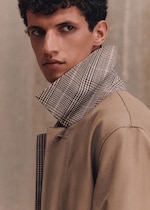 Placeholder for: A person with curly hair looking to the side, wearing a beige coat with a checkered collar turned up.
