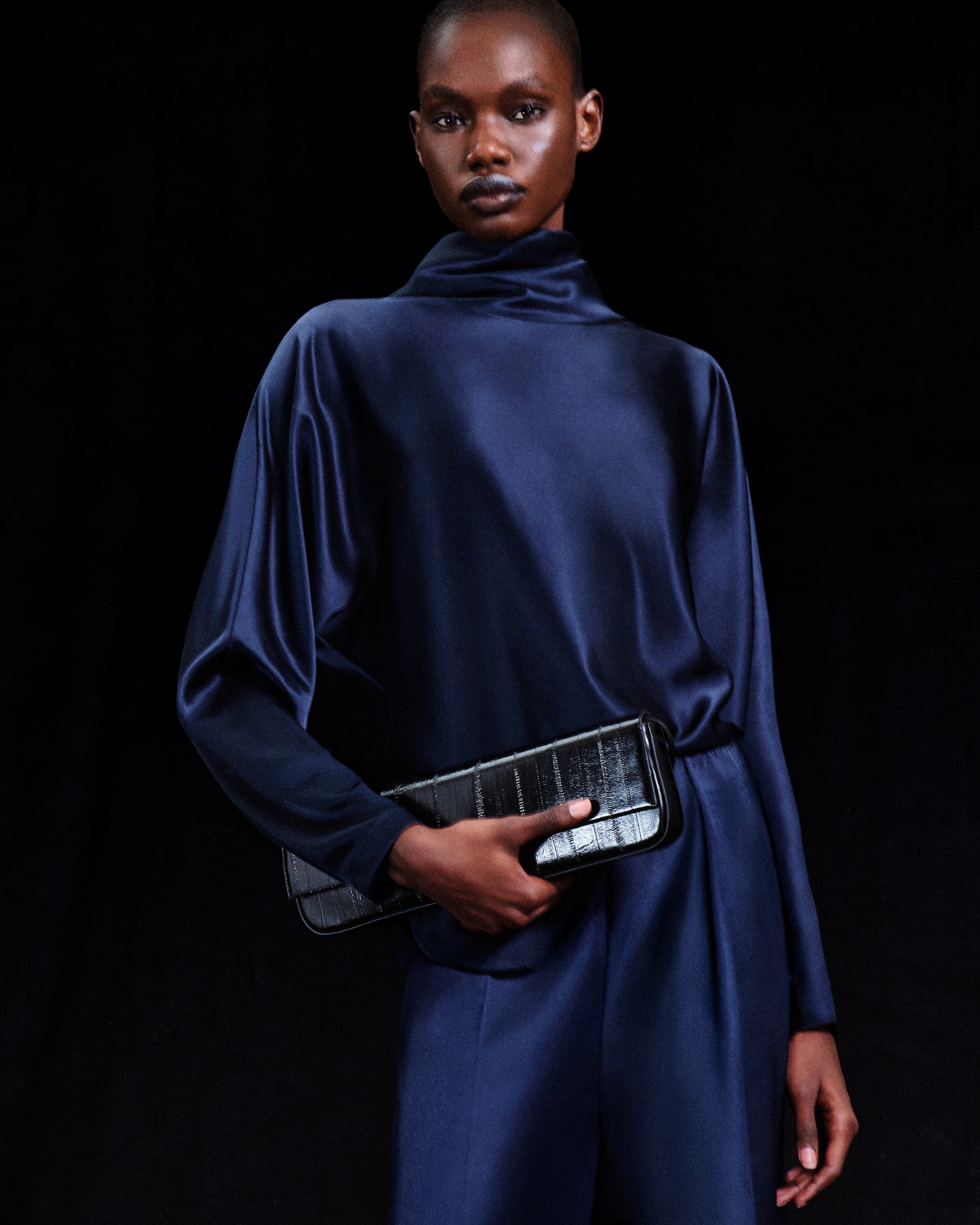 Person in a dark blue satin outfit, holding a black textured clutch, standing against a black background.