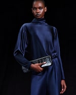 Placeholder for: Person in a dark blue satin outfit, holding a black textured clutch, standing against a black background.