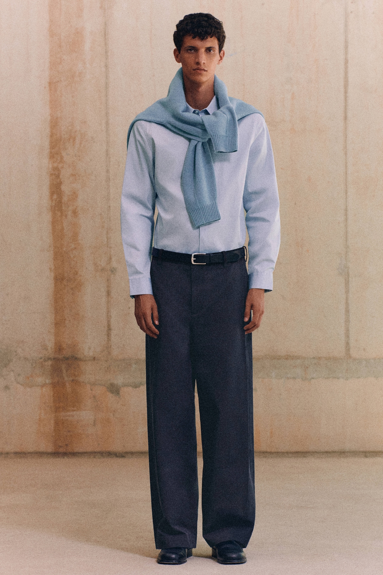 A person wears a light blue shirt and dark trousers, with a matching sweater draped over their shoulders against a simple beige background.