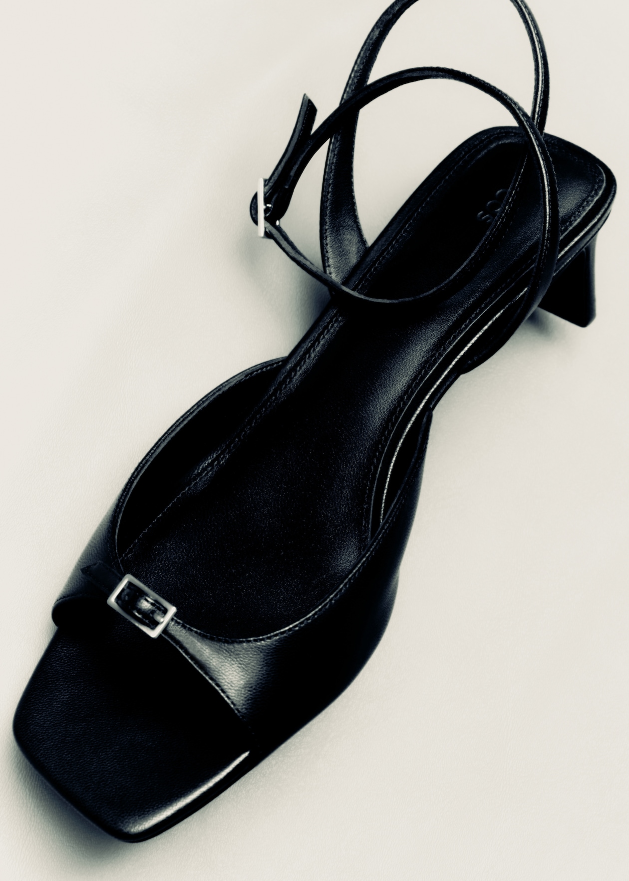 Black strappy sandal with a square toe and small buckle, featuring a low block heel on a light background.