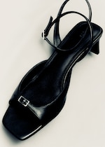Placeholder for: Black strappy sandal with a square toe and small buckle, featuring a low block heel on a light background.