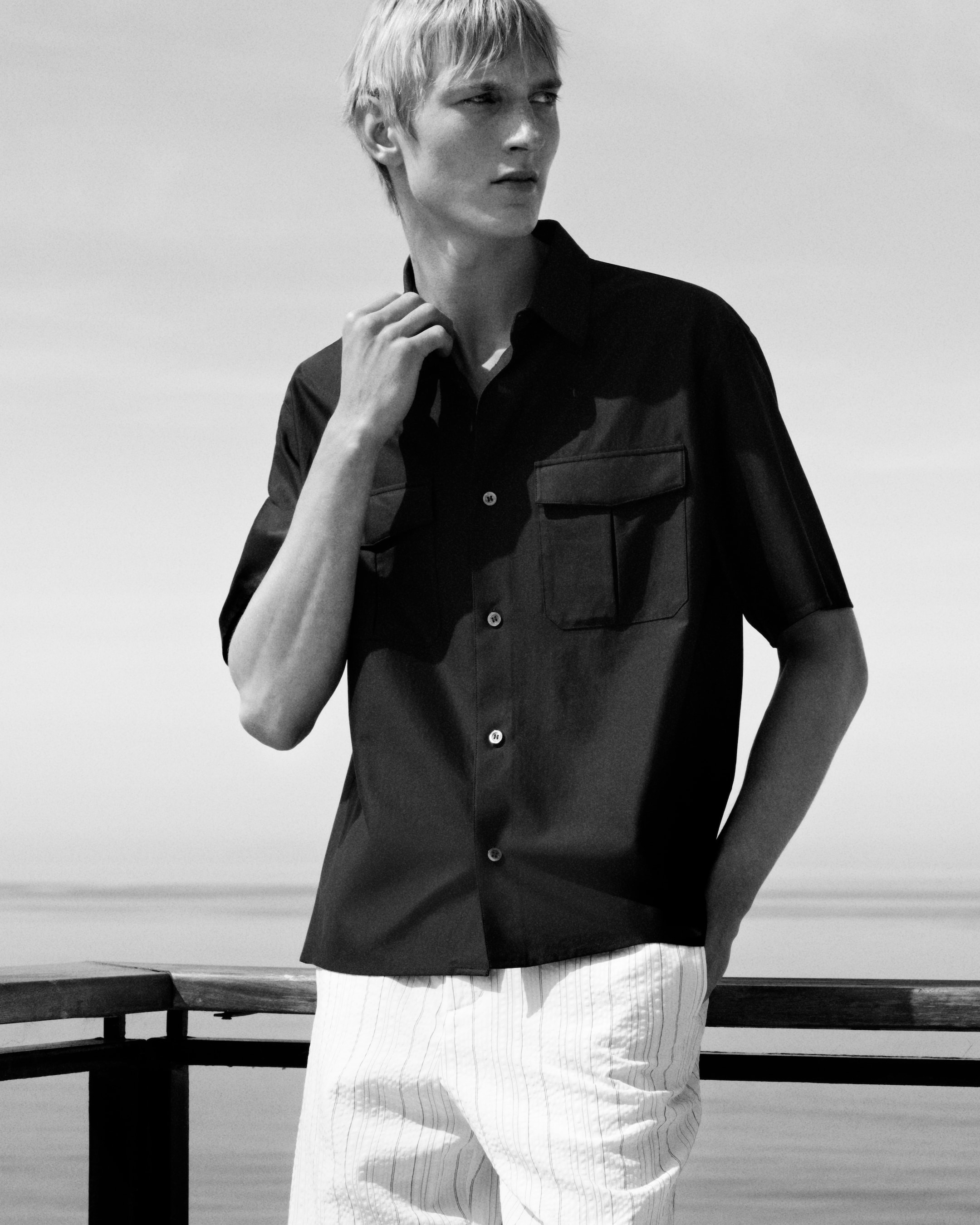 Black and white photo of a person in a dark shirt and light pants standing by a railing outdoors, gazing away thoughtfully.