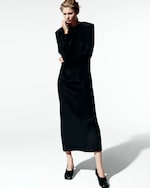 Placeholder for: A woman in a long, elegant black dress stands pensively against a plain, light background, with her hand on her chin.