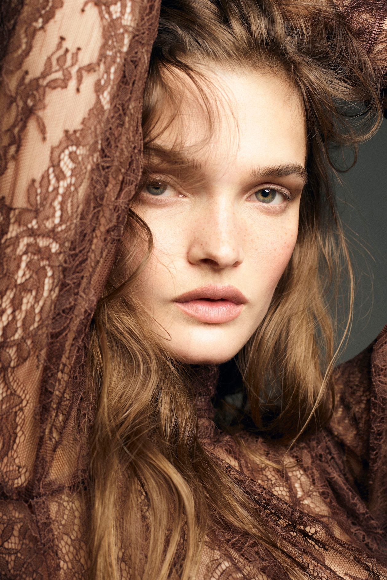Woman with long wavy hair and freckles, wearing a brown lace garment, gazes intensely at the camera, with arms raised behind her head.