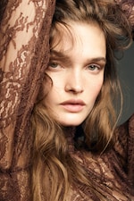 Placeholder for: Woman with long wavy hair and freckles, wearing a brown lace garment, gazes intensely at the camera, with arms raised behind her head.