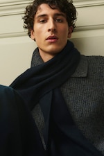 Placeholder for: A person with curly hair wearing a blue sweater and dark scarf, looking directly at the camera against a neutral background.