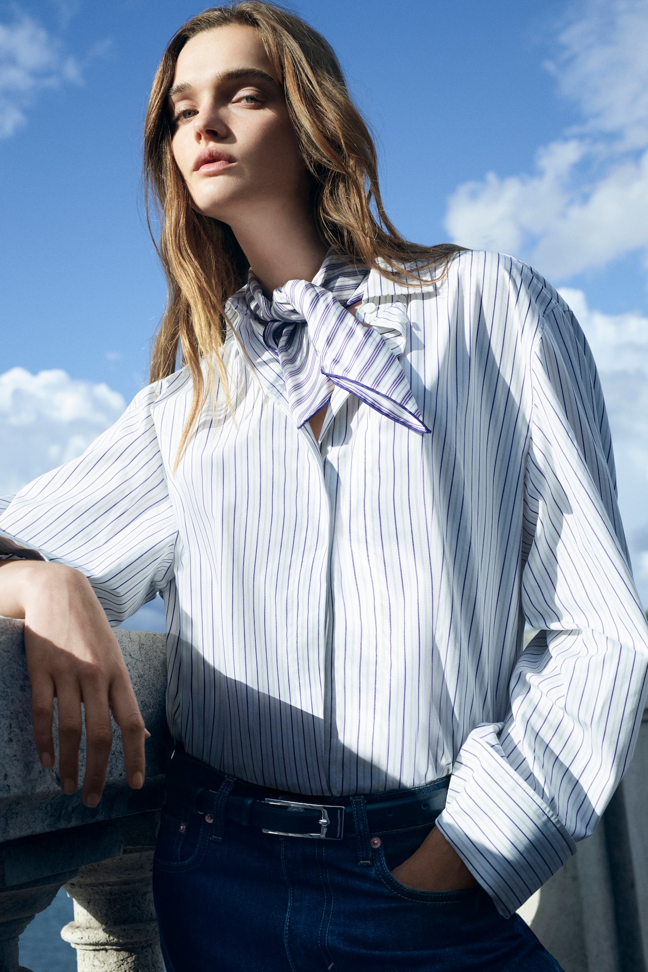 A person wearing a striped shirt and scarf stands confidently against a stone railing with a blue sky backdrop.