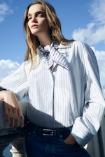 Placeholder for: A person wearing a striped shirt and scarf stands confidently against a stone railing with a blue sky backdrop.