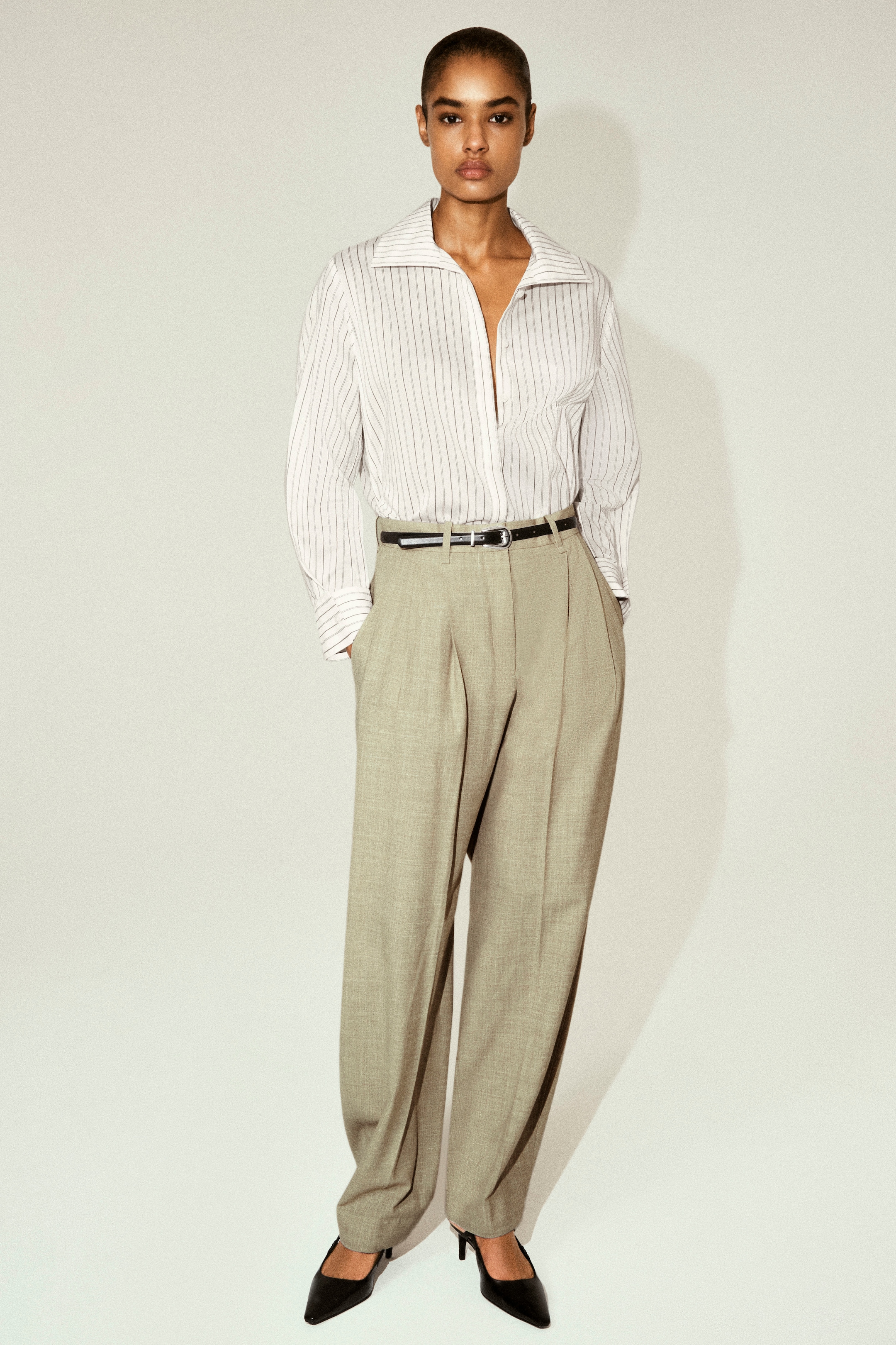 A person wearing a white striped shirt, beige pleated trousers, and black pointed heels stands against a neutral background, hands in pockets.