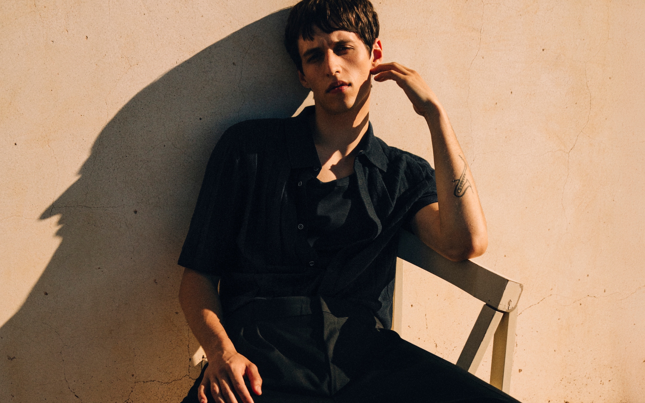 Person in black clothing and flip-flops sits on a chair in sunlight against a beige wall, casting a shadow on the tiled floor.