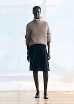 Placeholder for: A person stands indoors wearing a beige sweater, a dark pleated skirt, and black loafers, with soft light filtering through large windows.