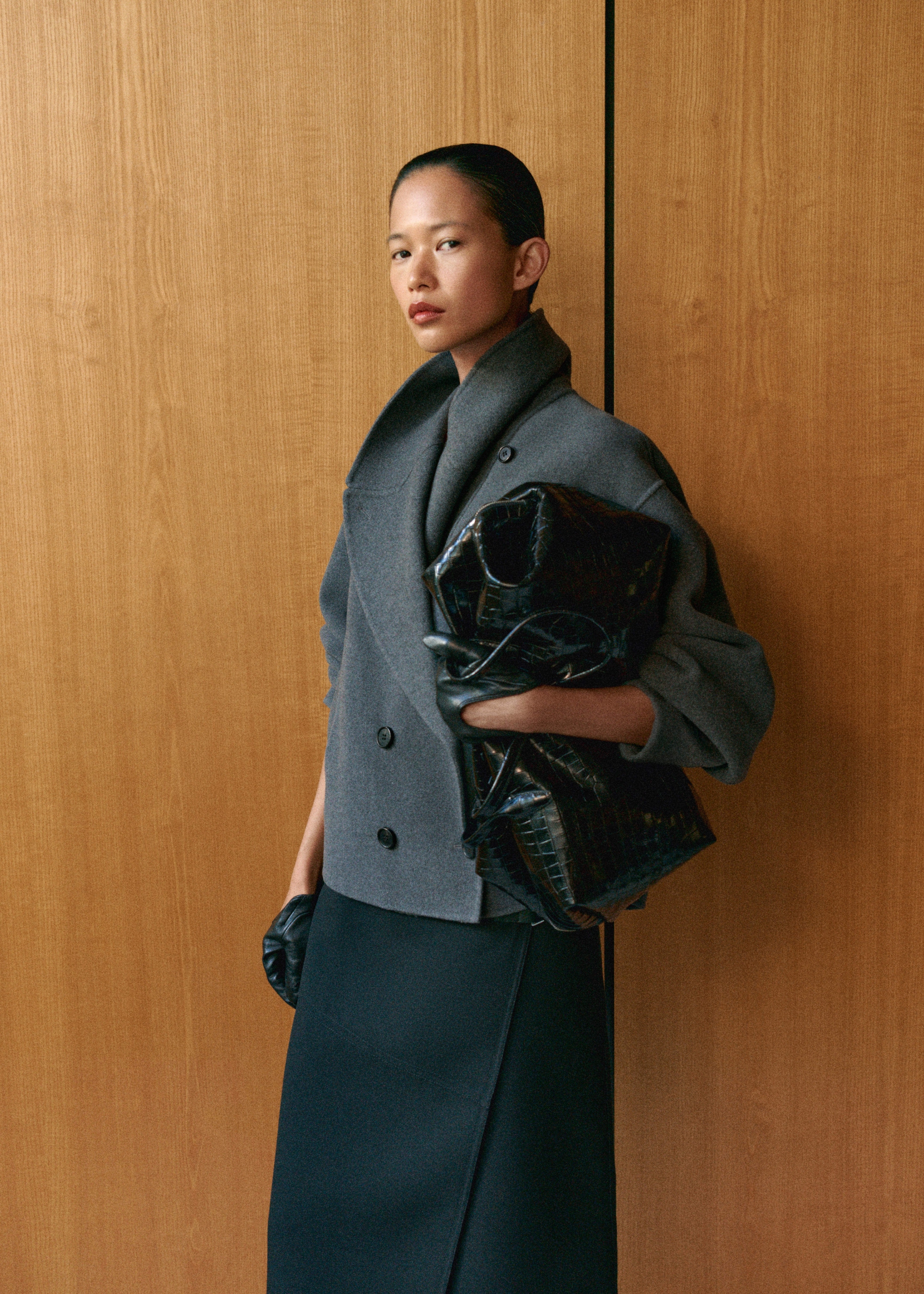 A person poses against a wooden background, wearing a gray coat and holding a black textured bag, with a calm and confident expression.