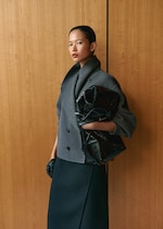 Placeholder for: A person poses against a wooden background, wearing a gray coat and holding a black textured bag, with a calm and confident expression.