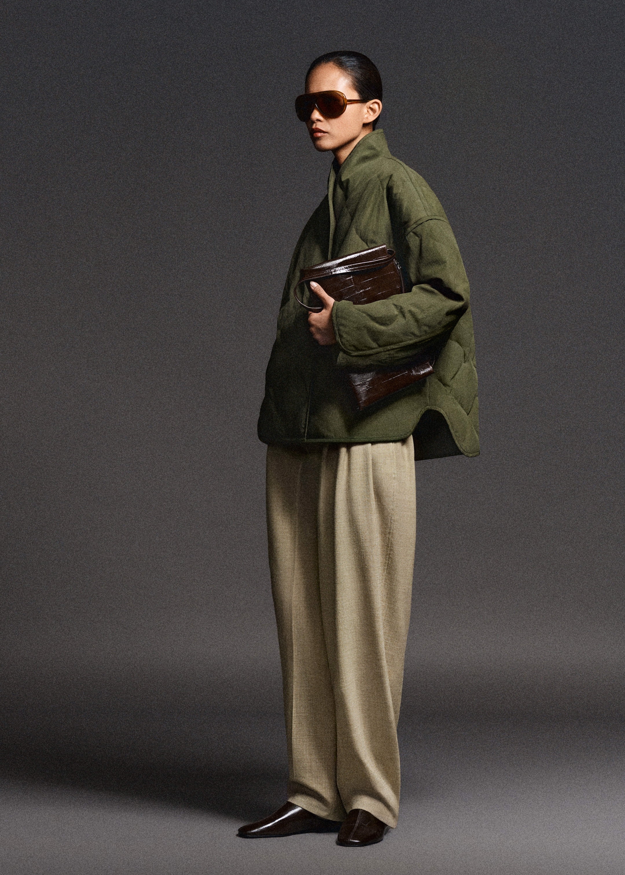 Person in oversized green jacket and beige trousers, holding a brown textured bag, wearing large sunglasses, standing against a gray background.