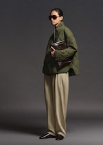 Placeholder for: Person in oversized green jacket and beige trousers, holding a brown textured bag, wearing large sunglasses, standing against a gray background.