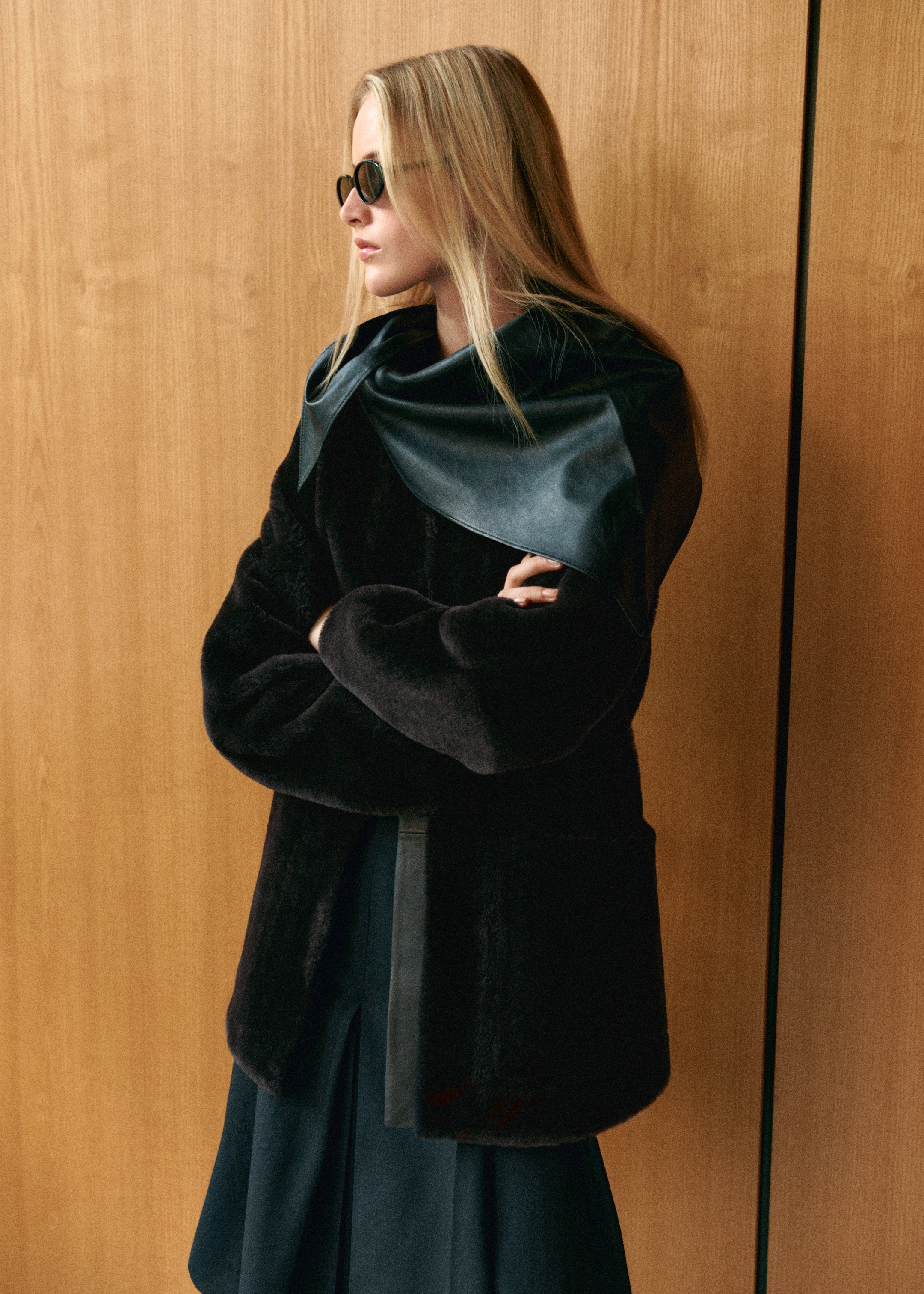 A person with long blonde hair, wearing sunglasses and a dark coat with a leather scarf, stands against a wooden background, looking sideways.