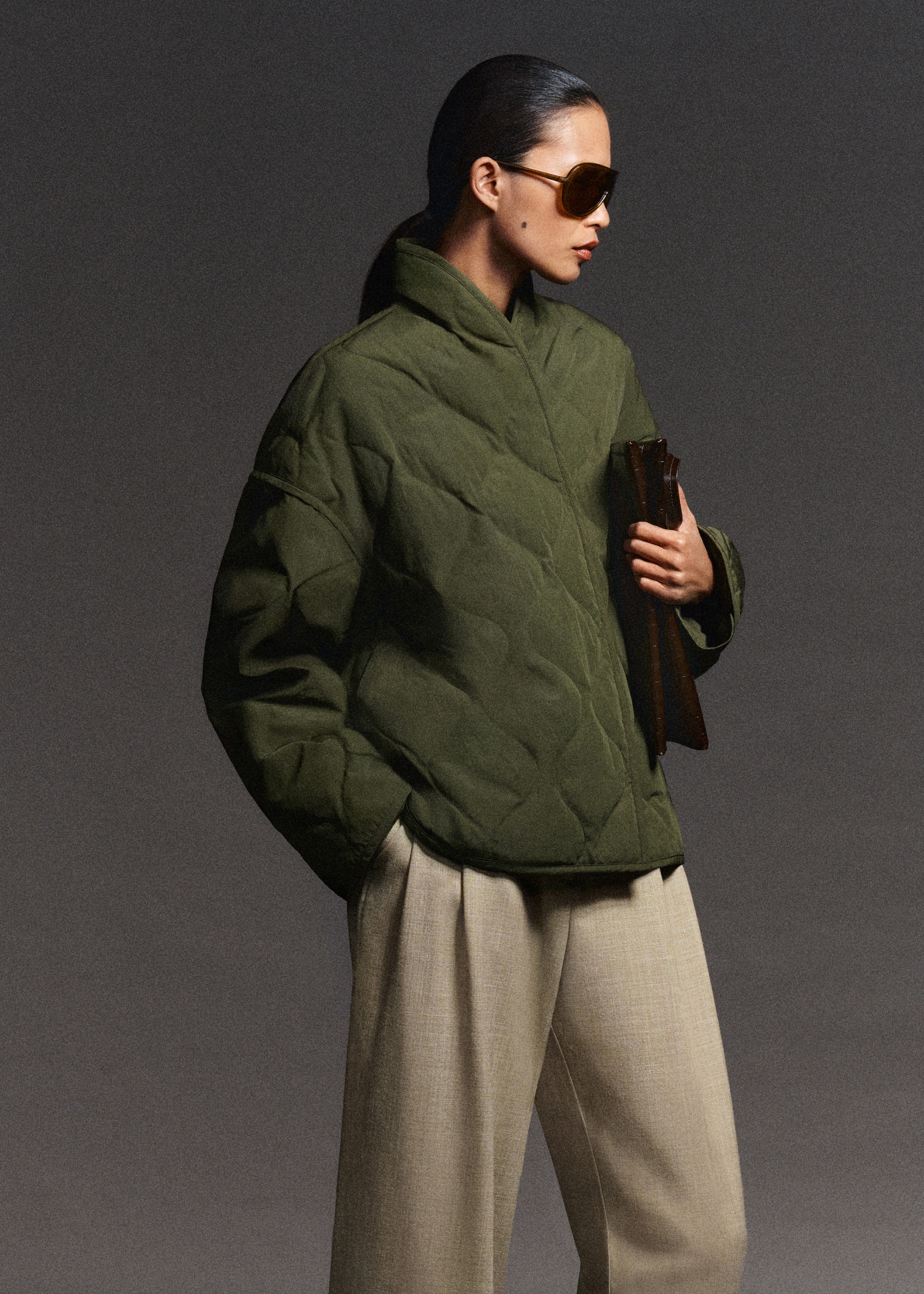 Person in a green quilted jacket and beige trousers, wearing sunglasses and holding a clutch, stands against a gray background.