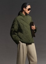 Placeholder for: Person in a green quilted jacket and beige trousers, wearing sunglasses and holding a clutch, stands against a gray background.
