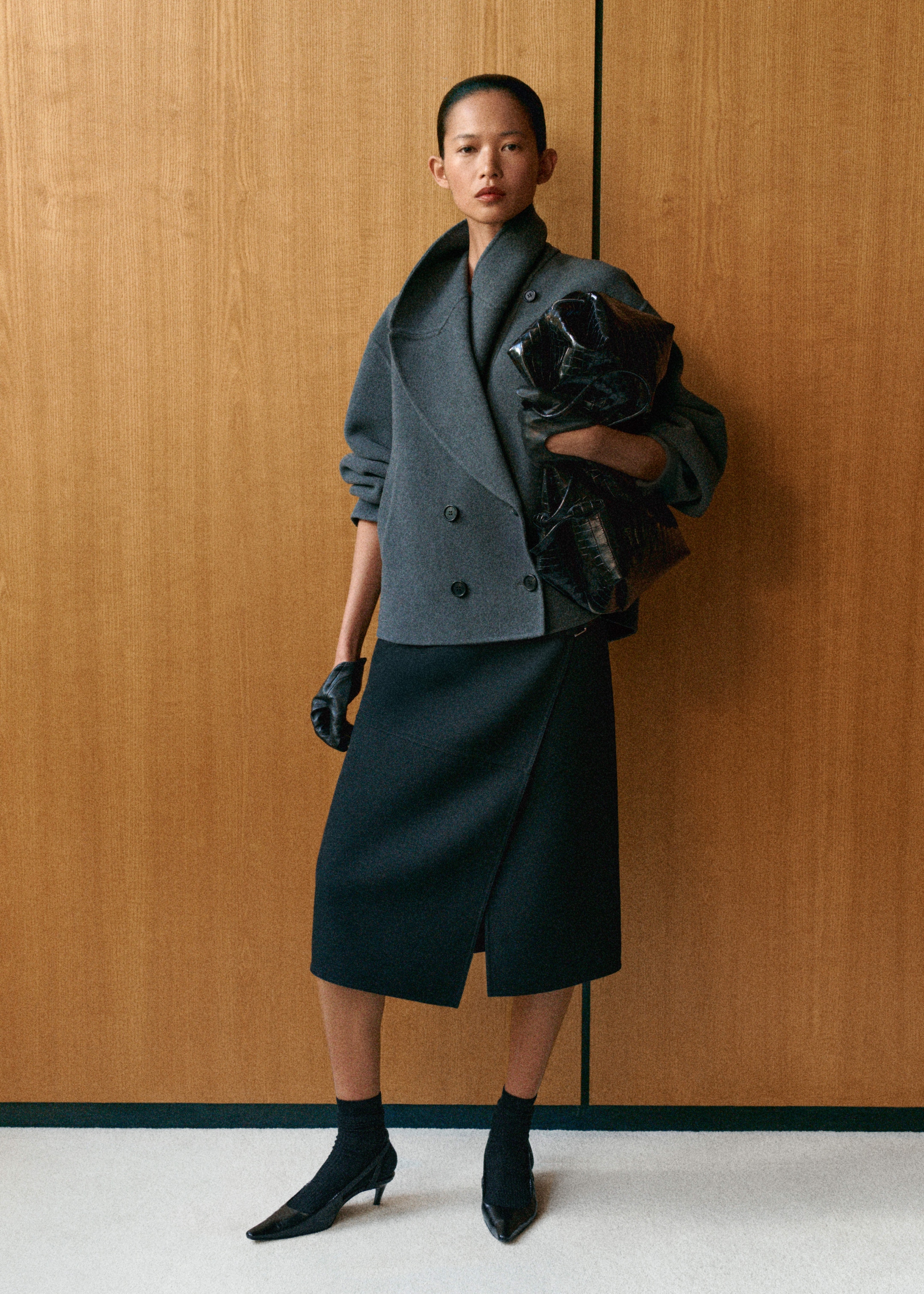 A person in a structured gray coat and black skirt, holding a glossy black bag, standing against a wooden background.