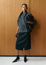 Placeholder for: A person in a structured gray coat and black skirt, holding a glossy black bag, standing against a wooden background.