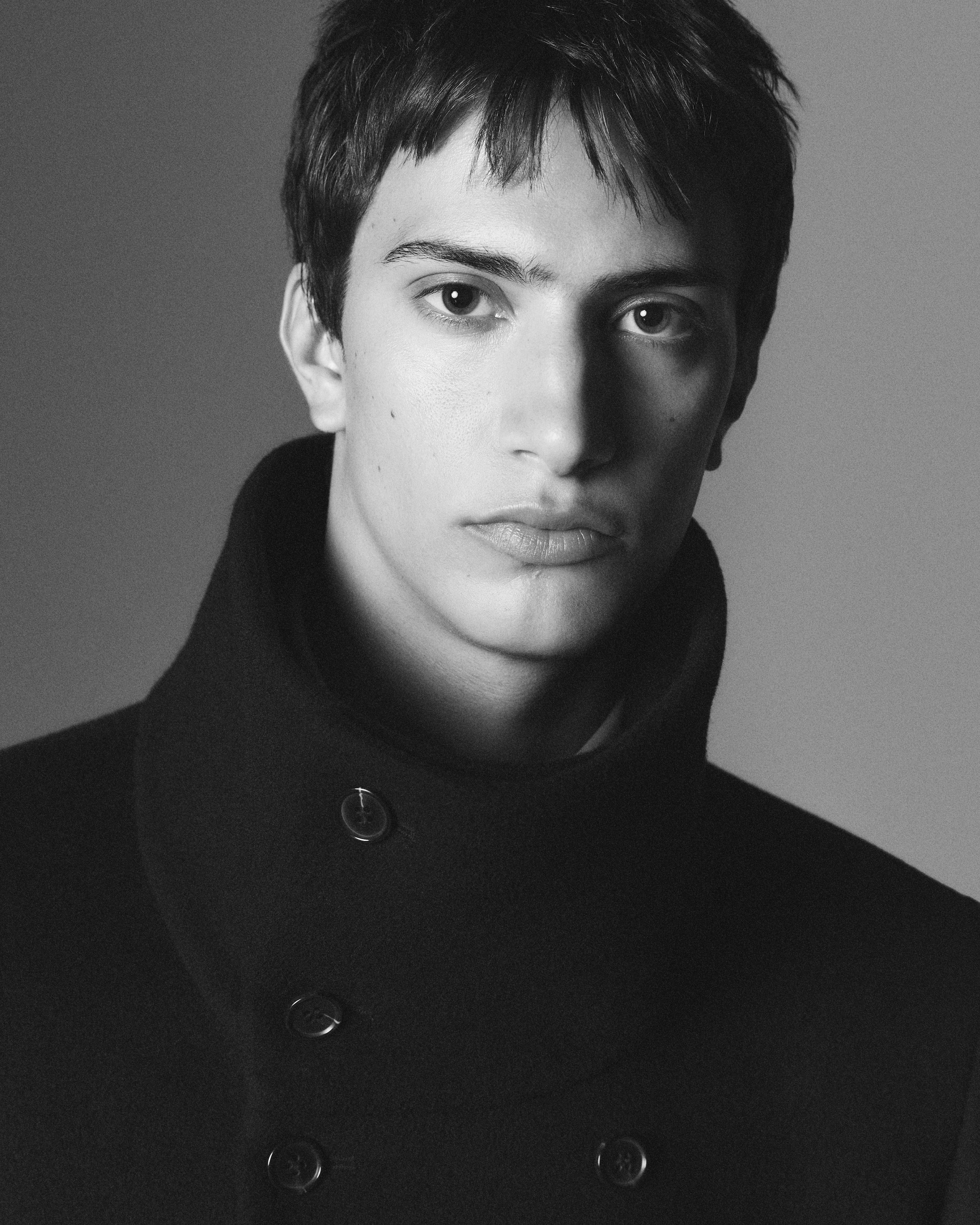 Black and white portrait of a person wearing a dark coat with a high collar, looking directly at the camera with a neutral expression.