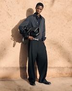 Placeholder for: Person in a dark, loose-fitting outfit stands against a textured beige wall, holding a large black bag. Shadows cast across the ground.