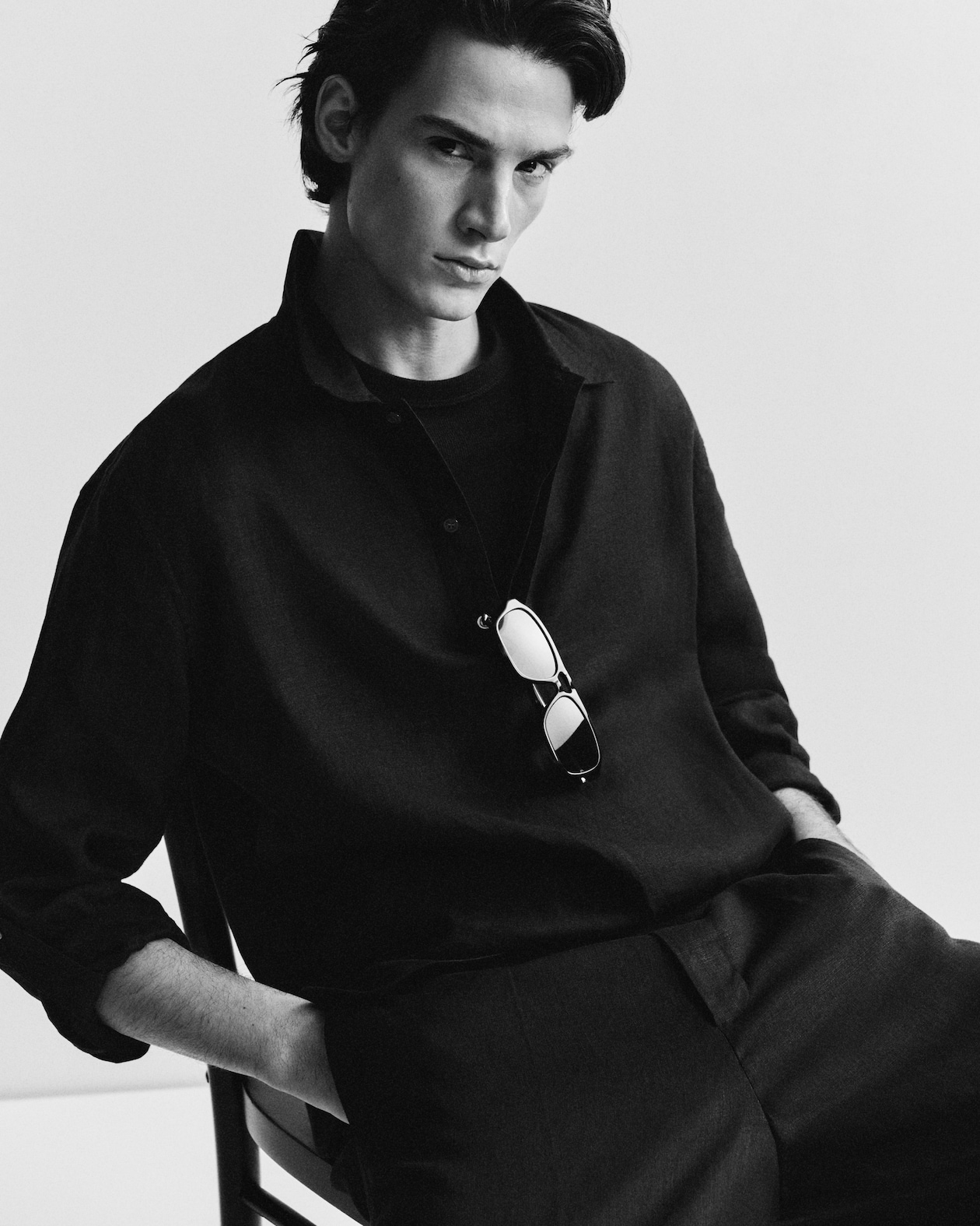 Black and white image of a person in a dark shirt and pants, sitting with hands in pockets. Sunglasses hang from the shirt collar.