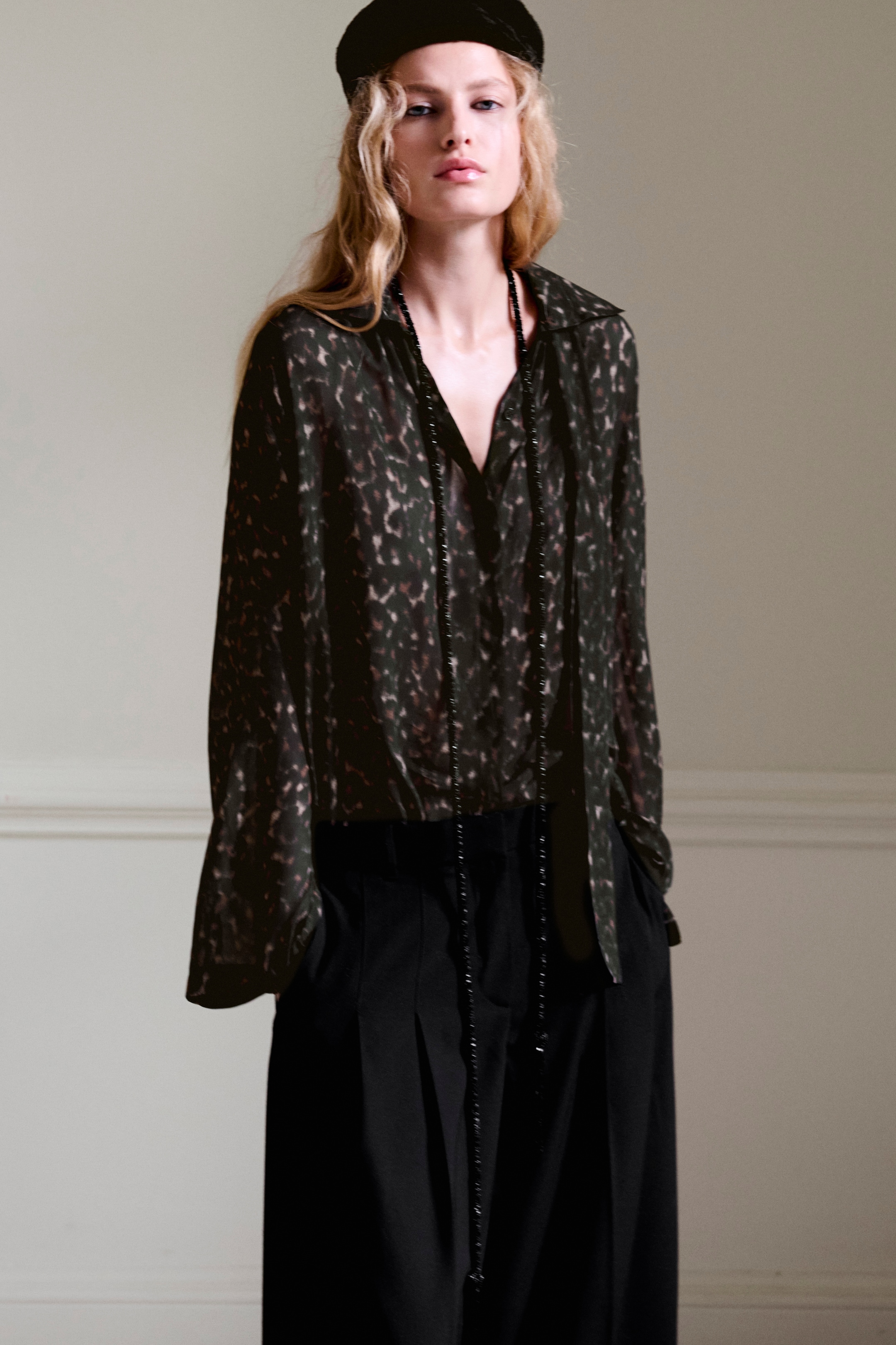 A person wearing a dark patterned blouse, black pants, long necklace, and a beret, standing against a neutral background.