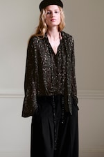 Placeholder for: A person wearing a dark patterned blouse, black pants, long necklace, and a beret, standing against a neutral background.