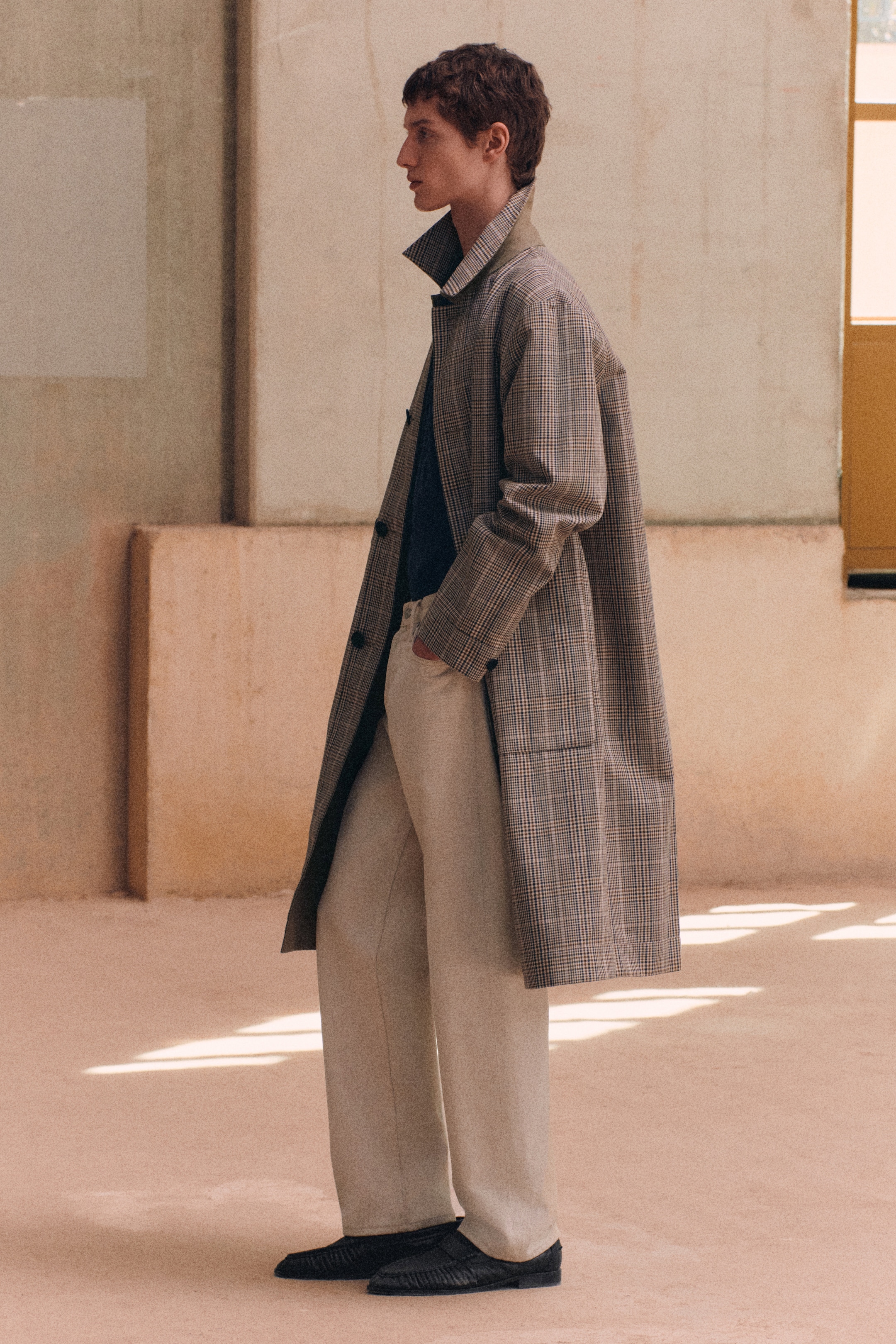A person in a plaid overcoat and light trousers stands in a minimalist room with muted tones and soft natural lighting.