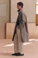 Placeholder for: A person in a plaid overcoat and light trousers stands in a minimalist room with muted tones and soft natural lighting.
