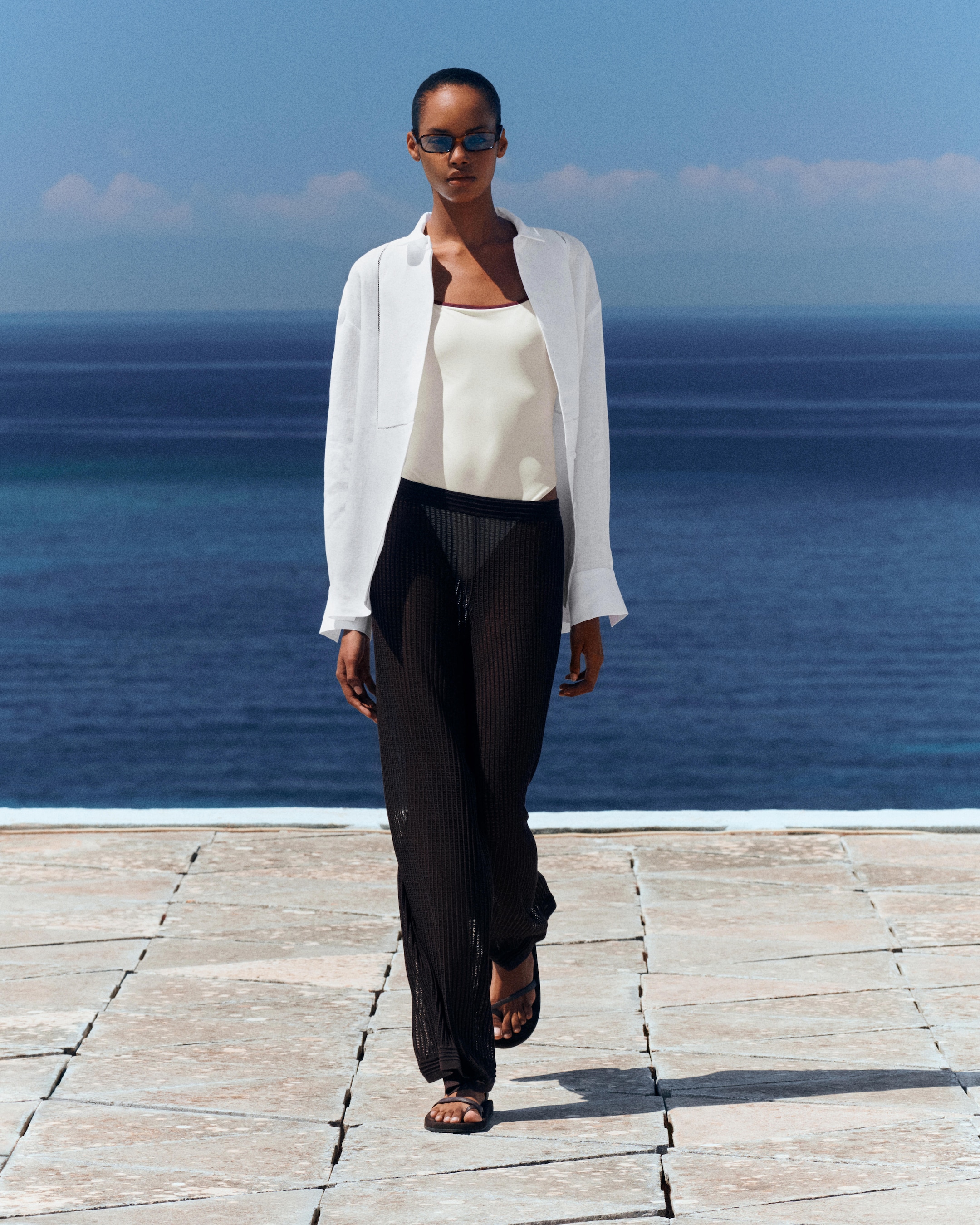 Woman in a white blazer and black pants walks confidently on a stone path by the sea, wearing sunglasses and sandals under a clear blue sky.