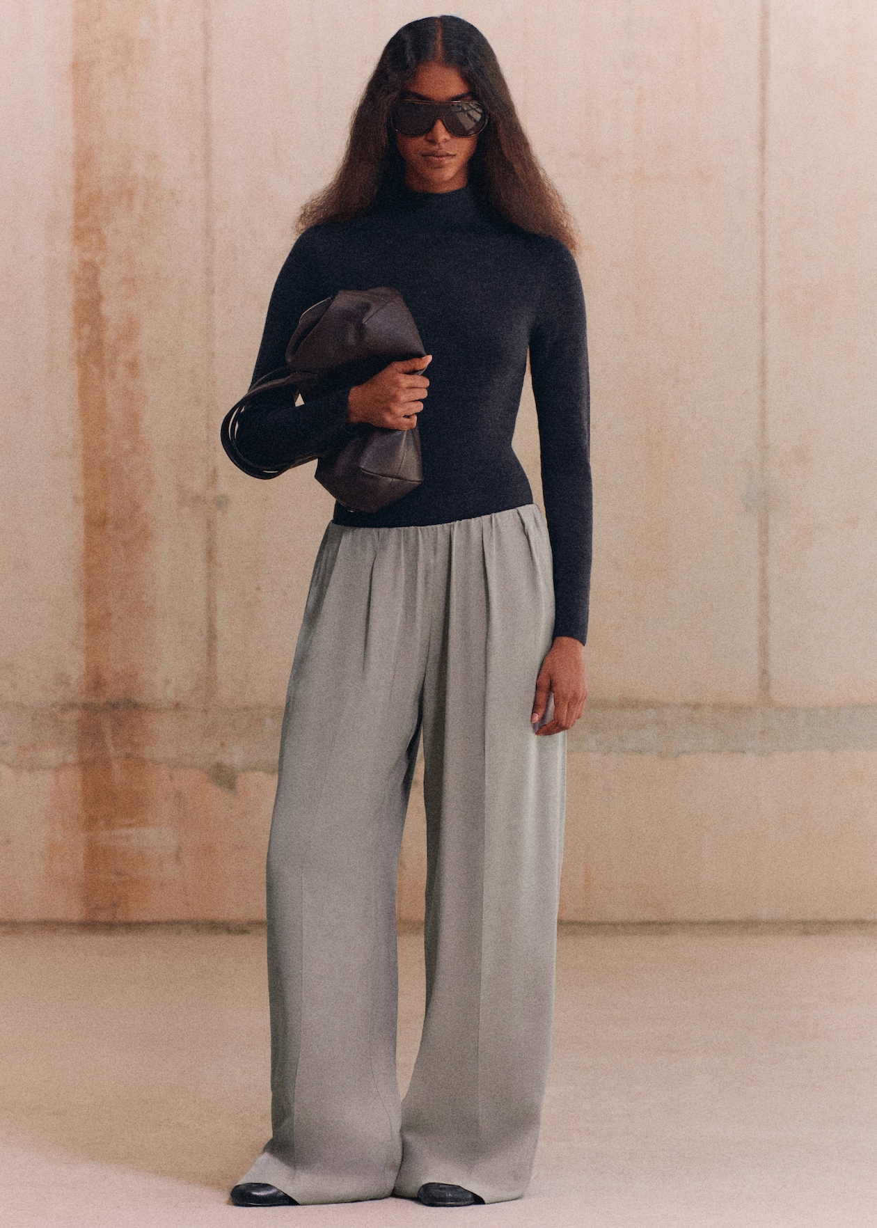 A person wearing sunglasses, a dark turtleneck, wide gray trousers, and holding a brown bag stands against a neutral background.