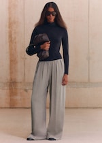 Placeholder for: A person wearing sunglasses, a dark turtleneck, wide gray trousers, and holding a brown bag stands against a neutral background.