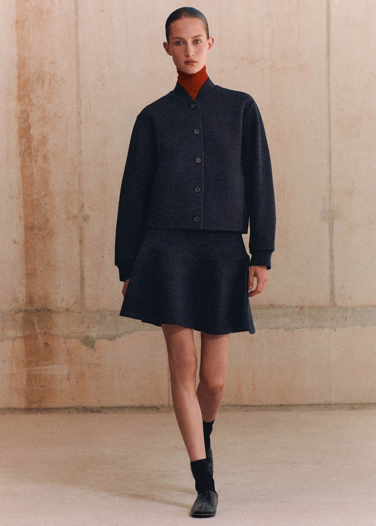 A person wearing a navy blue outfit, consisting of a buttoned jacket and skirt, with a red turtleneck, black socks, and shoes, standing in a minimalist room.