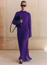 Placeholder for: A person in a flowing purple dress and sunglasses walks confidently, holding a black handbag against a neutral background.