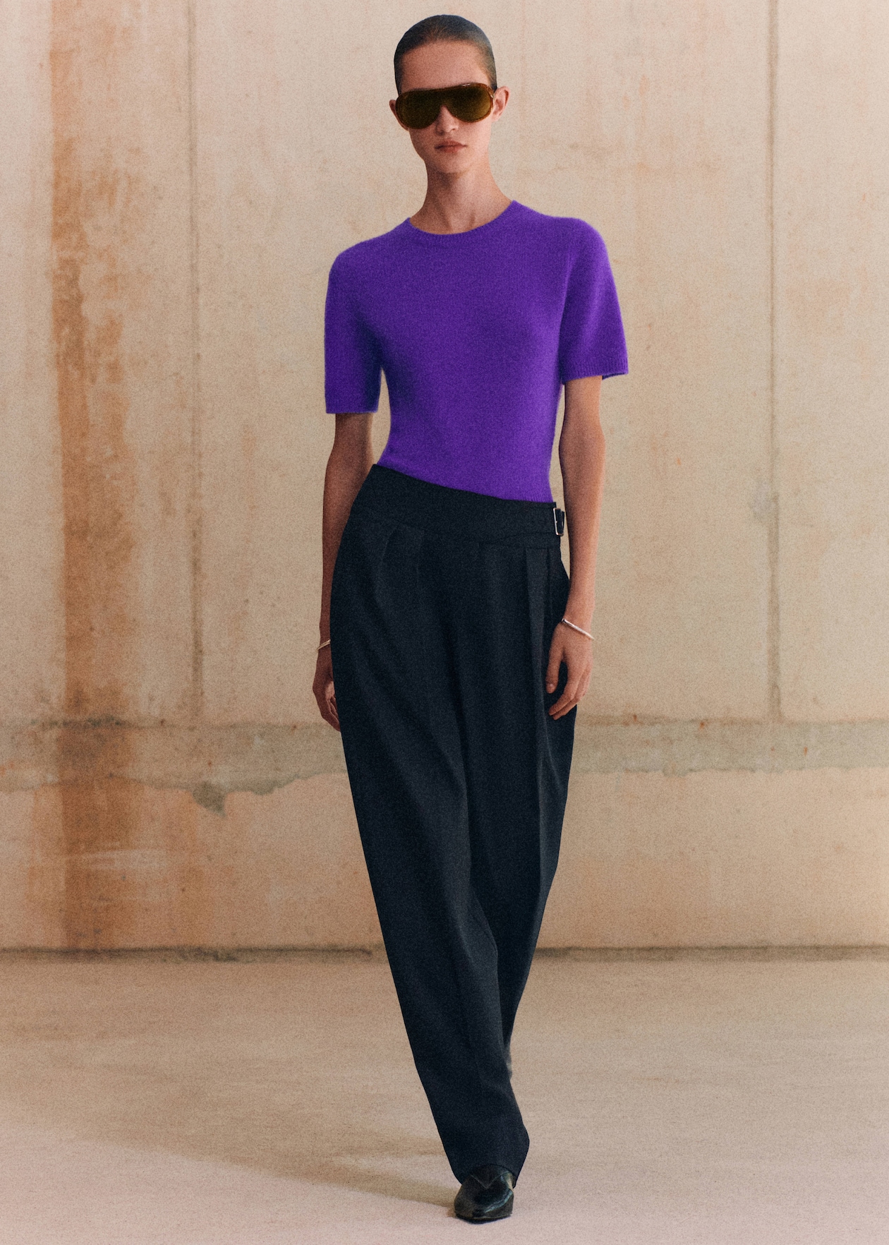 Person wearing a purple top, black pants, and large sunglasses, standing against a beige textured wall.