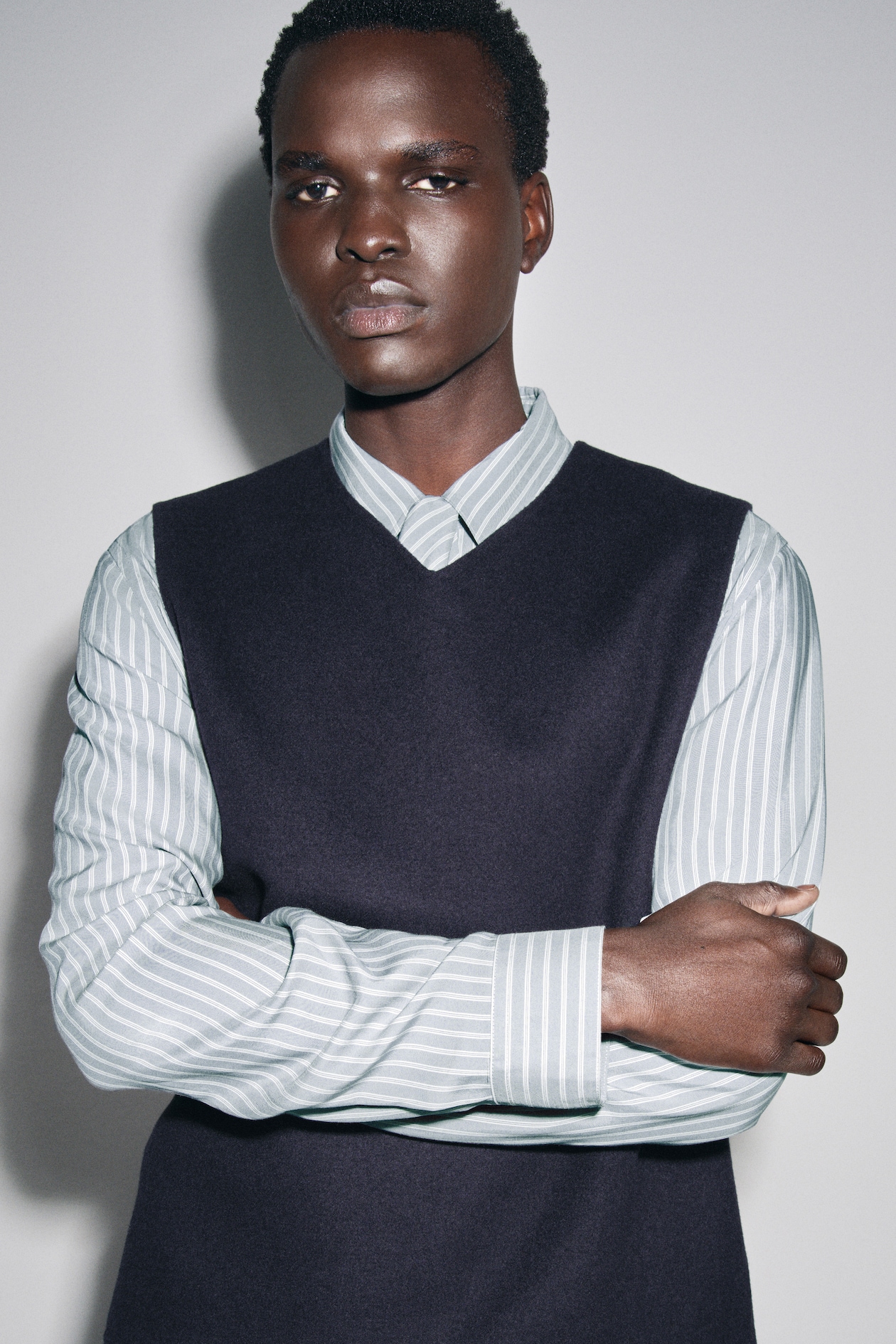 Person with short hair wearing a striped button-up and dark sleeveless sweater, standing with arms crossed against a light background.