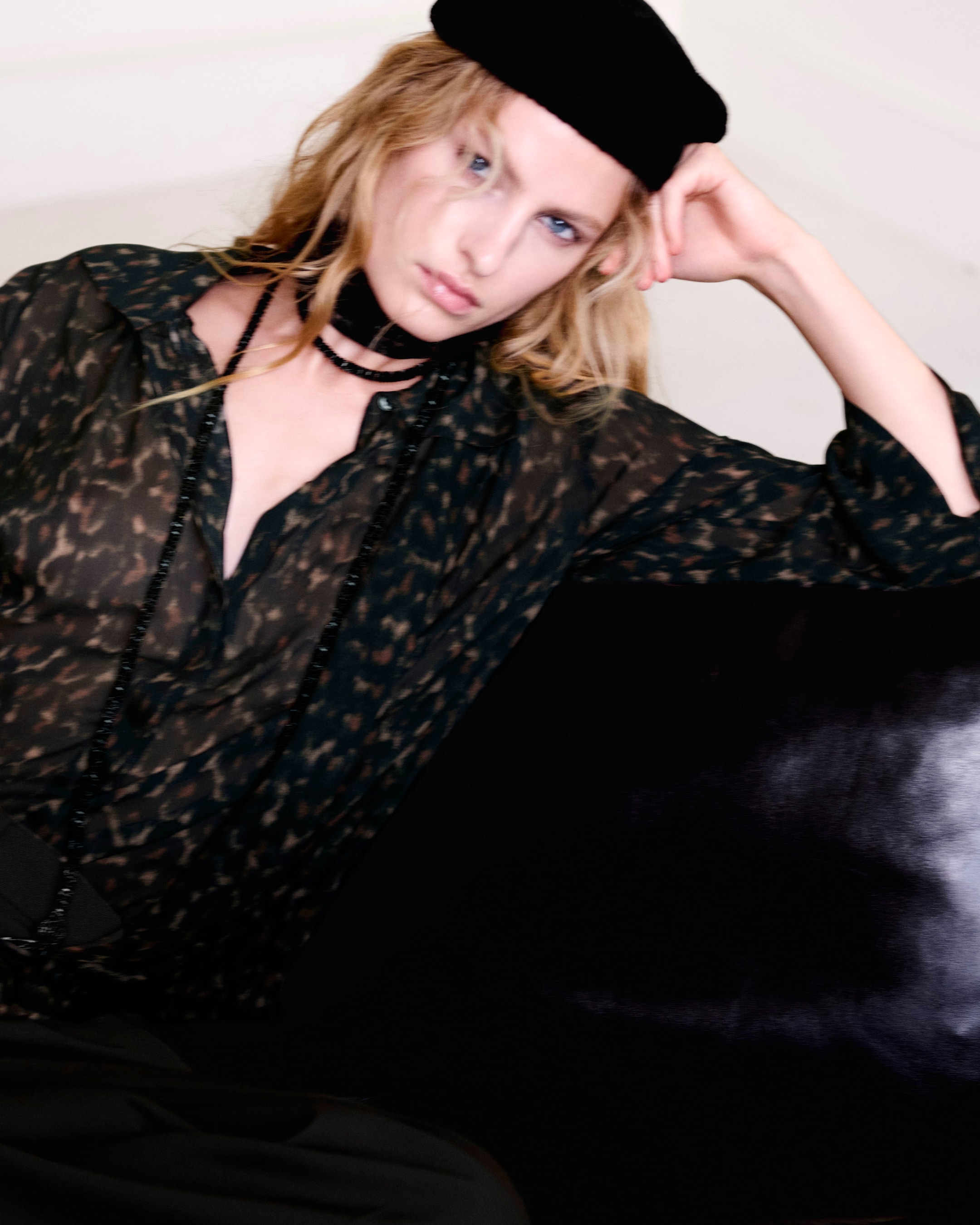 Person wearing a black beret and patterned blouse, sitting with a relaxed pose, resting their head on one hand with a neutral expression.