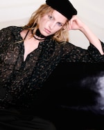 Placeholder for: Person with blonde hair wearing a black beret and patterned shirt, resting on a dark couch, gazing intently at the camera.