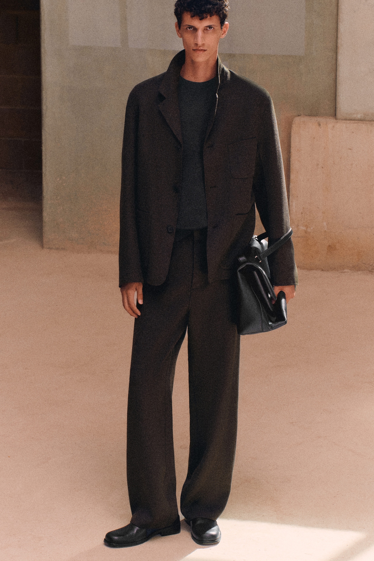A person in a dark, loose-fitting suit stands against a minimalistic beige wall, holding a black bag.
